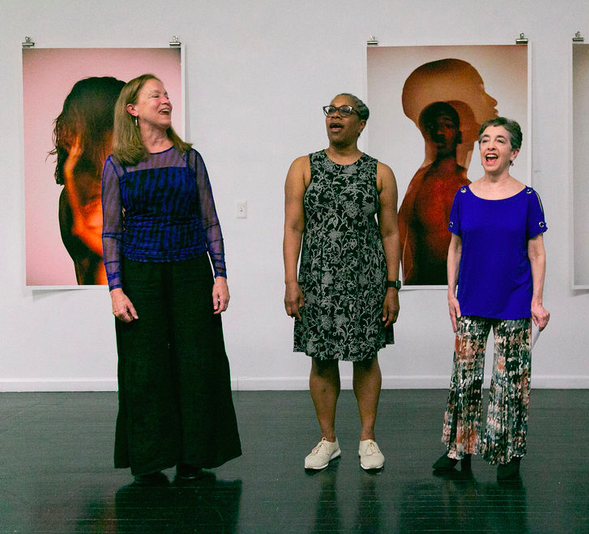 Three women sing Songs From the Hill by Meredith Monk against a background of torso portraits by Whitney Browne